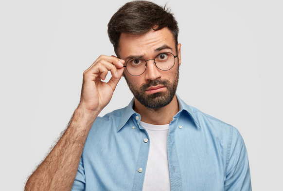 Attractive unshaven male looks curiously through glasses, keeps hand on rim